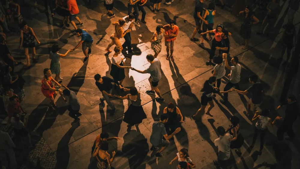 Overhead view of around thirty couples swing dancing on a paved square at night, long shadows across the stones